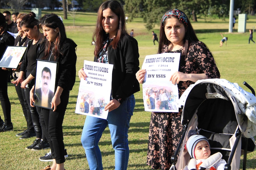 Ezidi women stand in park holding posters about the Ezidi genocide at a memorial in Toowoomba.