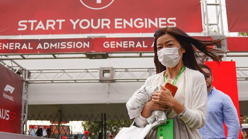 A woman wearing a face mask walks away from a sign at the gates of the track reading 'start your engines'.