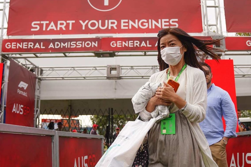 A woman wearing a face mask walks away from a sign at the gates of the track reading 'start your engines'.