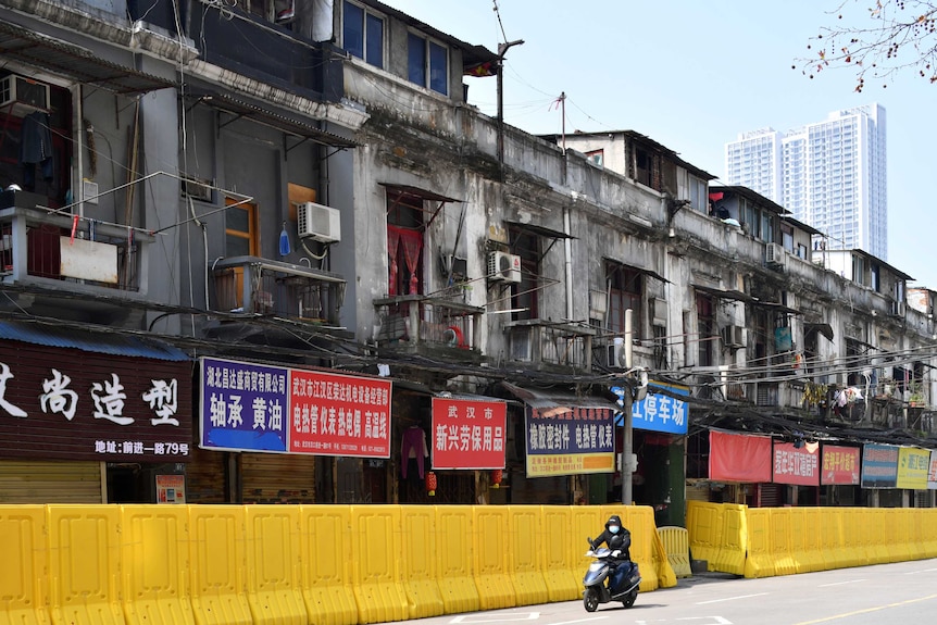 A man in a face mask rides a scooter past a row of apartments blocked by a yellow plastic barricade