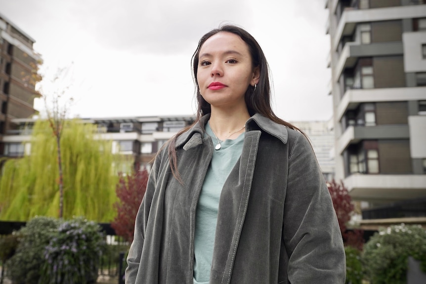 A young woman in a grey coat poses for the camera on an overcast day.