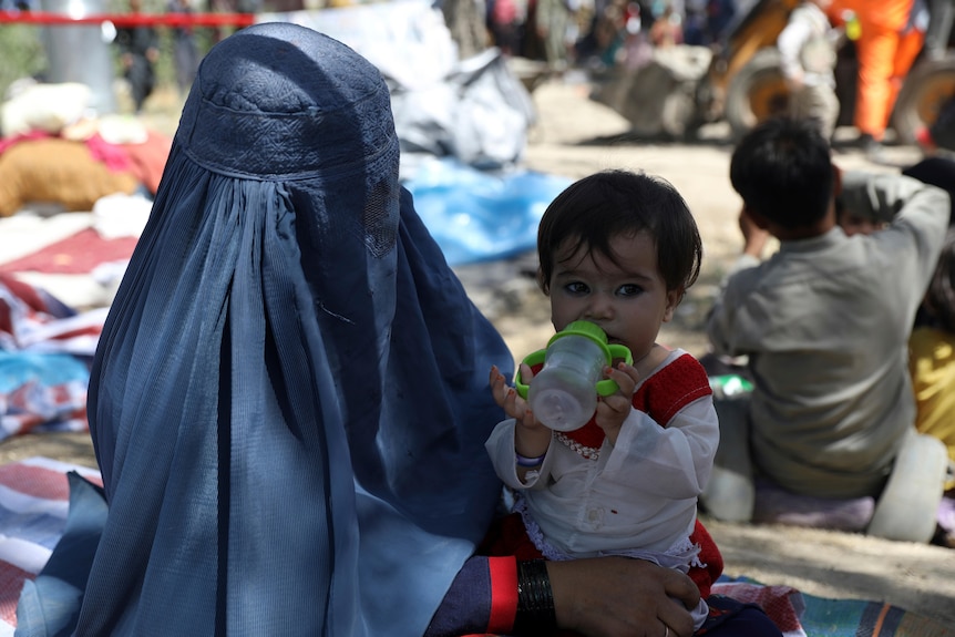 Woman wearing a blue burqa holds a young child drinking from a bottle. 
