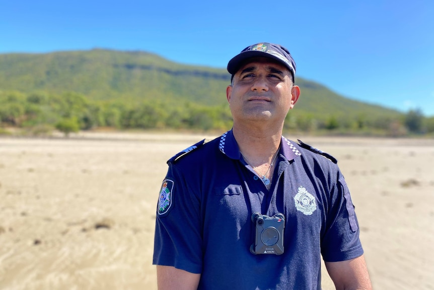 A police officer stands on a white beach with mountains in the background. It is a sunny day.