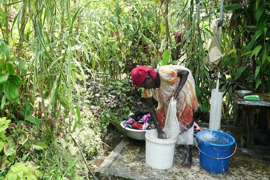 A woman standing over a bucket in an outside laundry, washing clothes and dishes.