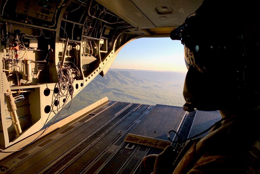 An Army crewman wearing a helmet looks out of a Chinook helicopter at a North Queensland landscape.
