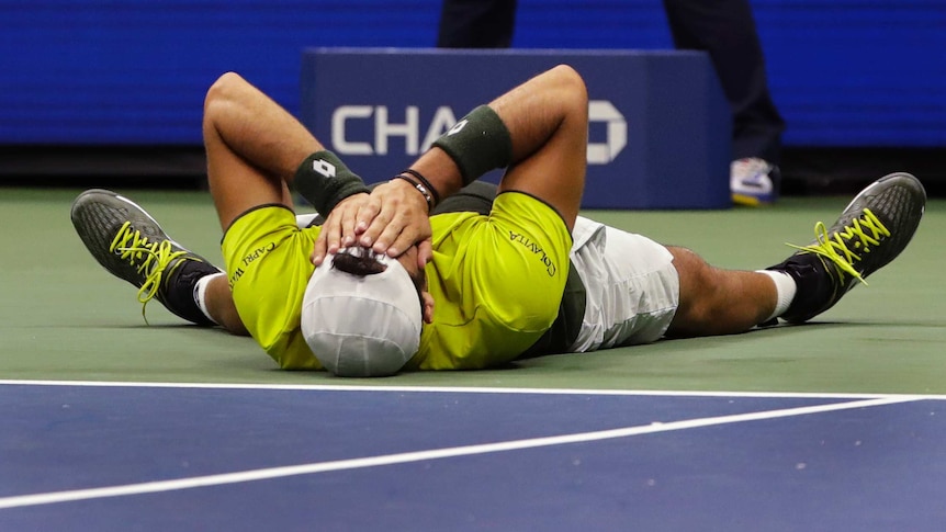 A male tennis player lies on the court with his hands covering his face as he celebrates winning a match.