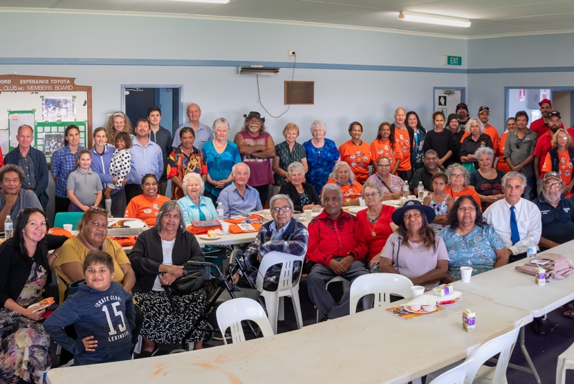 A group photo of dozens of people, many of them Indigenous and brightly dressed, in a community hall-style room.