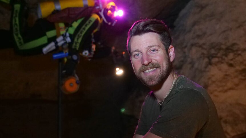 Ryan Kaczkowski crouches in a dark cave with multi-coloured lights and equipment in the background.