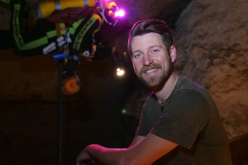 Ryan Kaczkowski crouches in a dark cave with multi-coloured lights and equipment in the background.