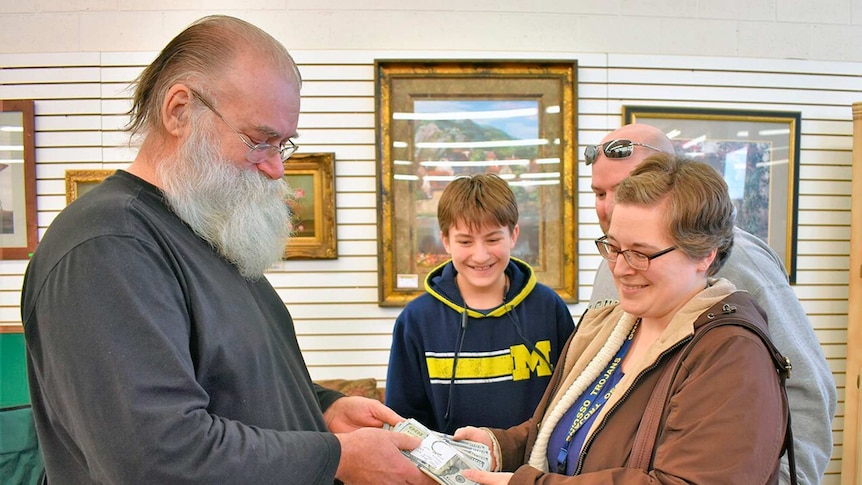 An elderly, bearded man hands stacks of cash to a woman and her family.