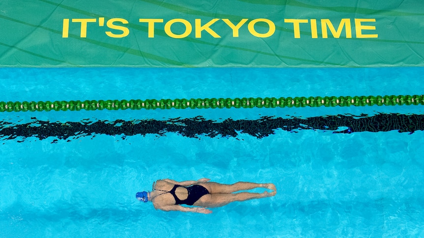A swimmer is underwater, seen from above, warming up at the Australian Olympic trials under a sign reading "It's Tokyo time".