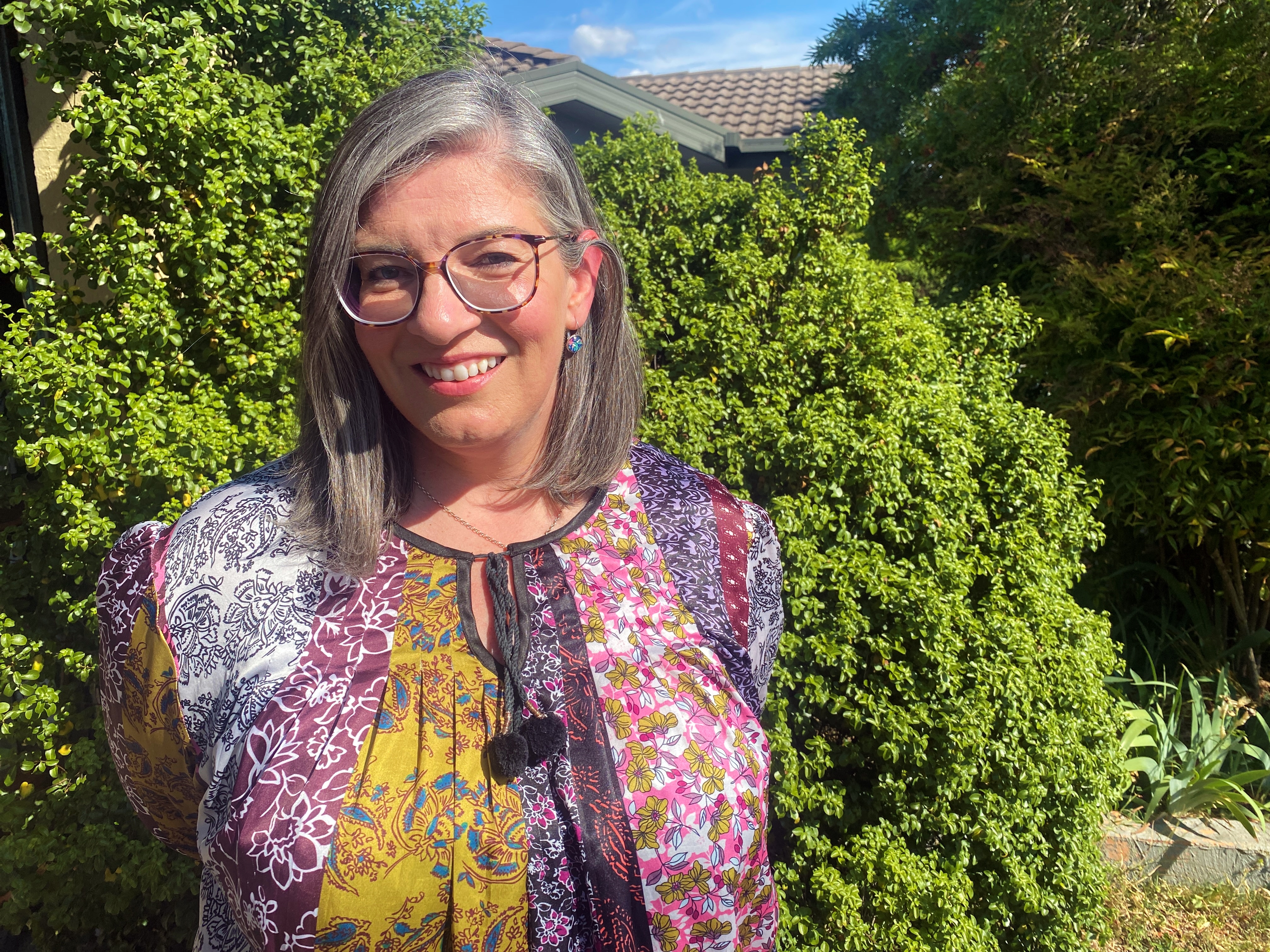 A woman with glasses smiles while standing in her front yard.