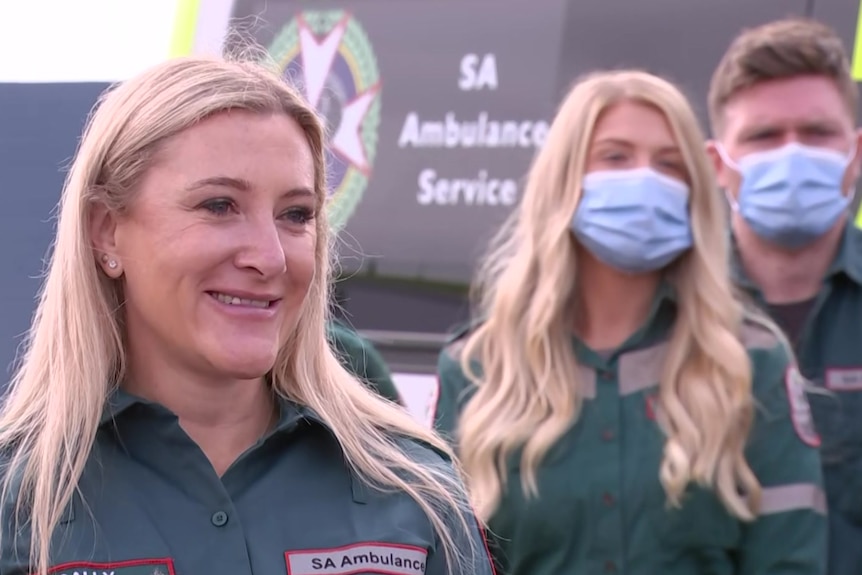A smiling woman wearing a green paramedic uniform, behind her are a woman and man wearing blue masks and green uniforms