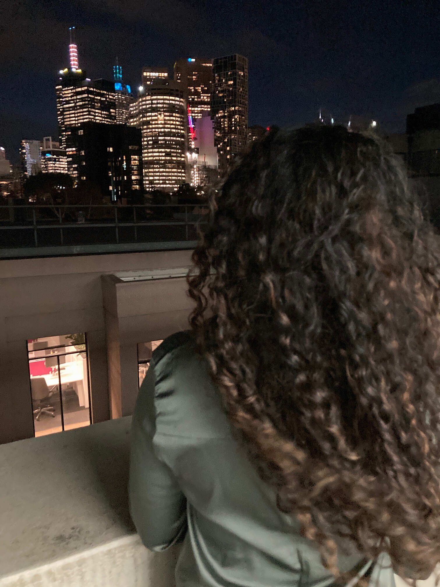 The back of a woman's head as she looks at the skyline of the city at night.