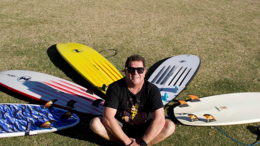 A man sits on the ground surrounded by surfboards in a fan shape.