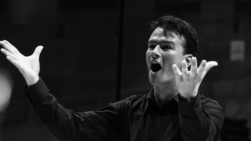 A black and white photo of a young man in a black shirt with his arms raised and mouth open singing.