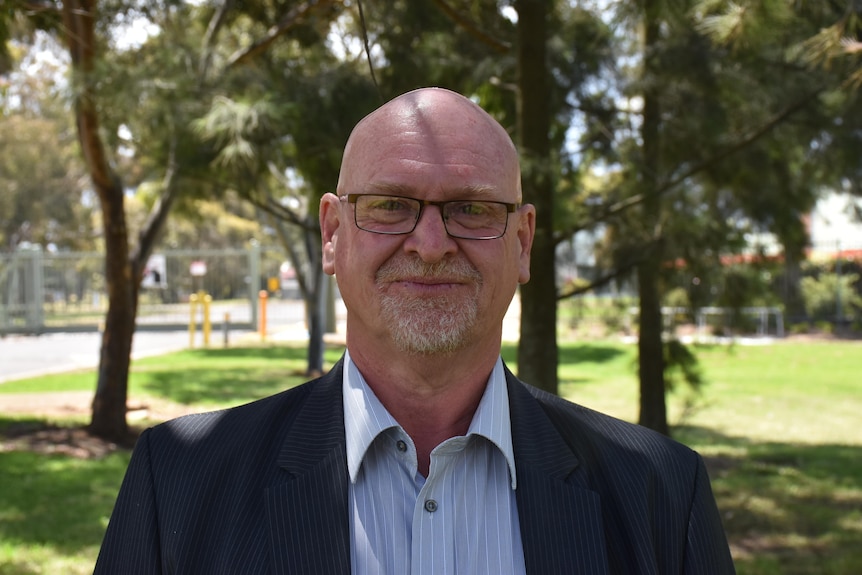 Peter Mayall stands beneath some trees beside a suburban road, in a shirt and jacket wearing glasses.