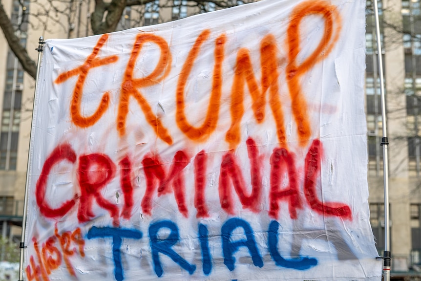 A banner says 'Trump criminal trial'. It is held up outside a courthouse.