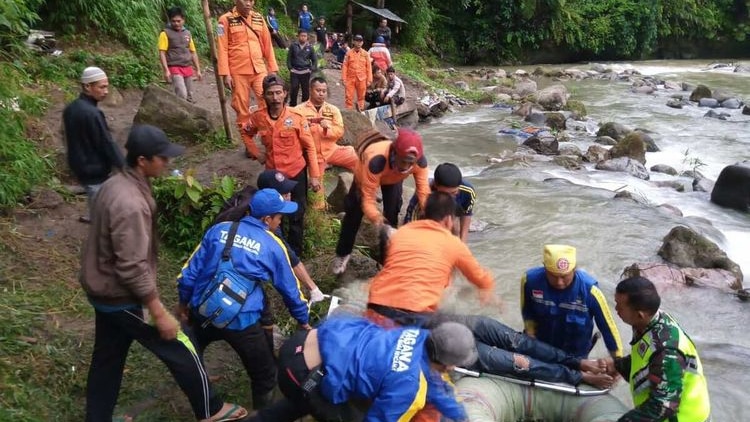 Looking down at a riverbank next to fast-flowing rapids, a line of people in orange place a body on a metal stretcher.