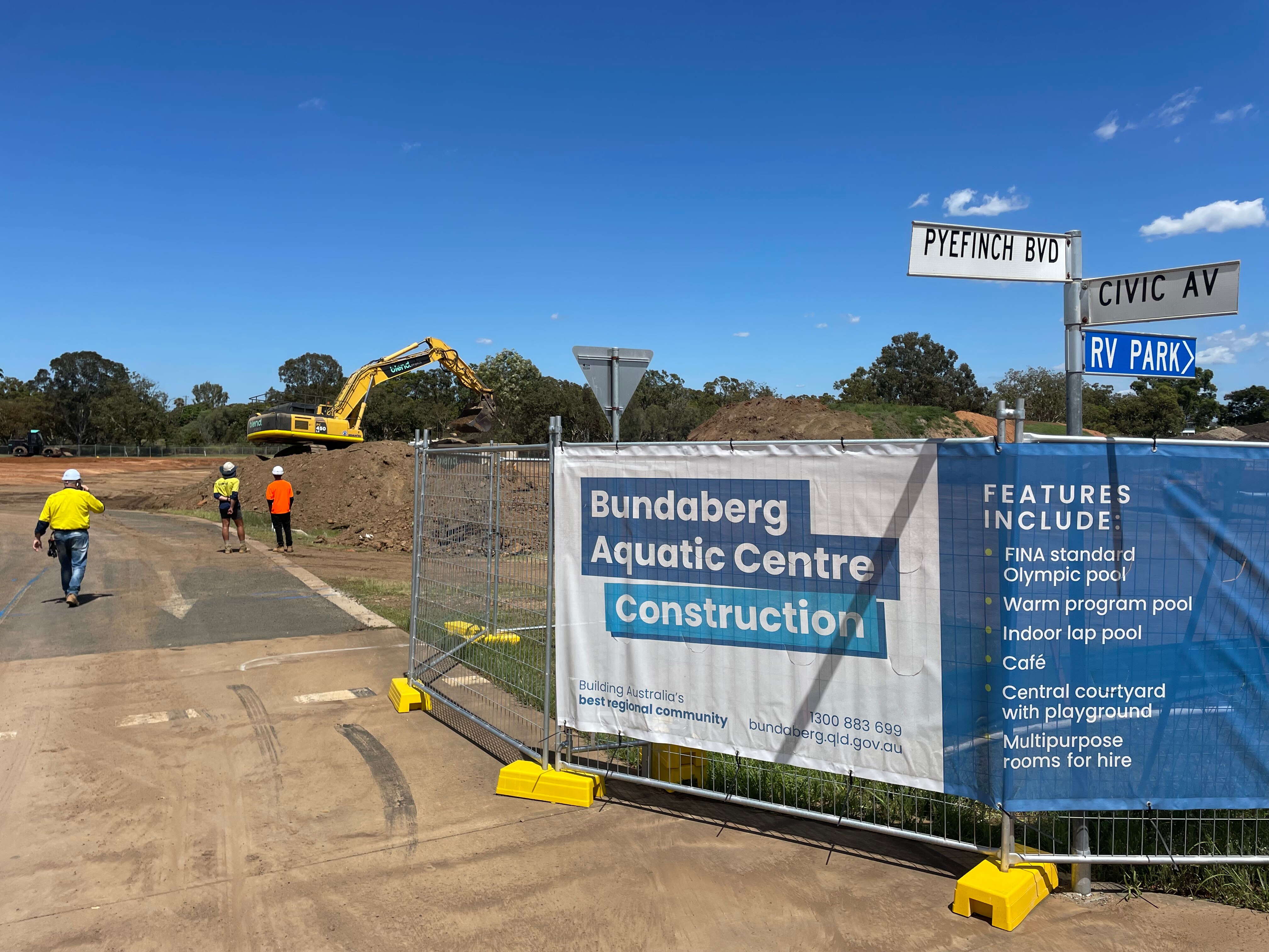 Construction fences stand in the foreground with workers in high-visibility shirts and an excvator in the backgrou 