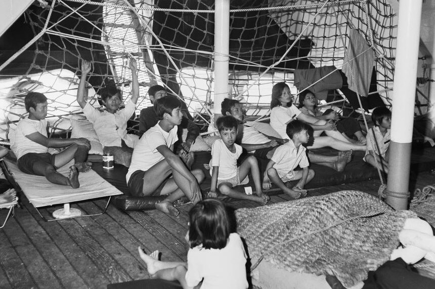 A large group of Vietnamese adults and children sit on mats on the floor of a ship's deck