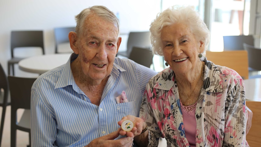 An elderly couple sit smiling at the camera, holding out a small gold medallion.