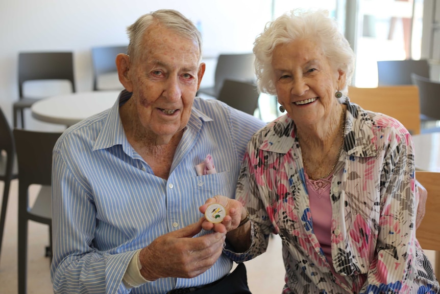 An elderly couple sit smiling at the camera, holding out a small gold medallion.