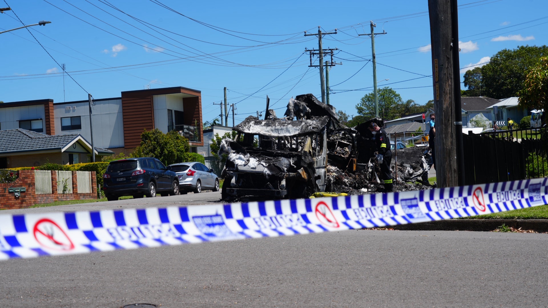 Blue and white police tape in the foreground, a burnt, blackened vehicle on the road in the background.