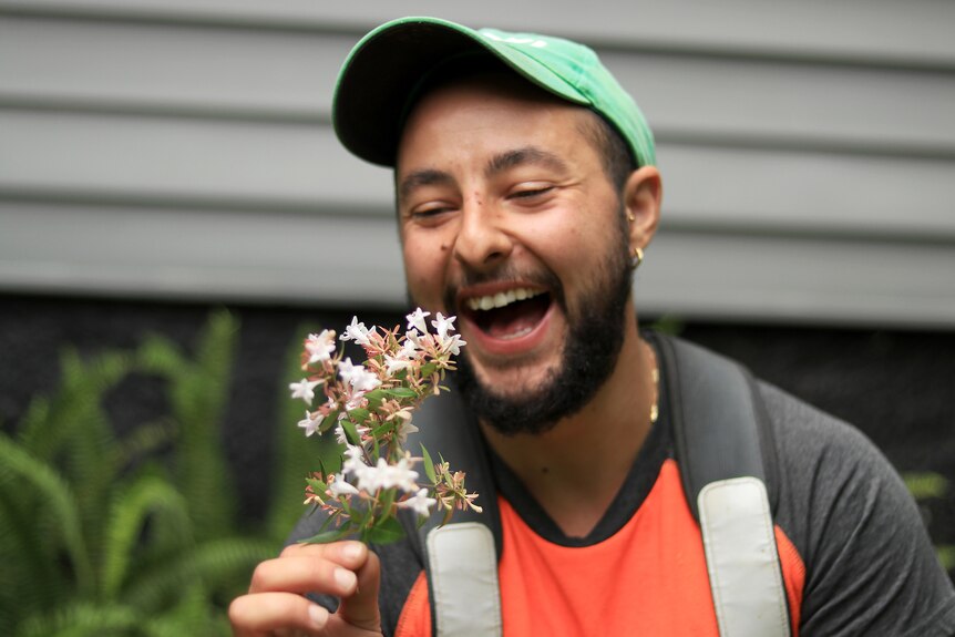 A laughing man holds some white flowers and laughs, wearing a green cap and orange and grey t-shirt.