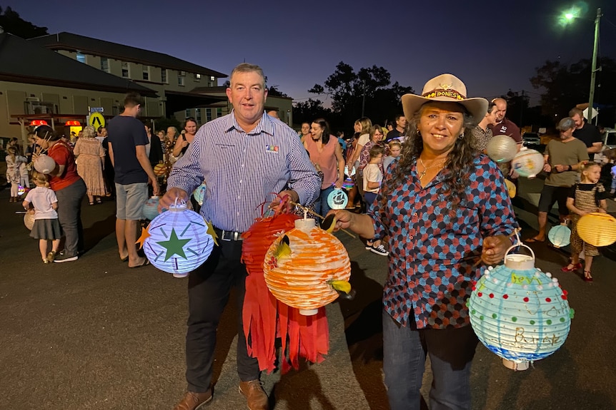 A man and an Indigenous woman hold paper lanterns in front of a crowd of townspeople on a street at nighttime.