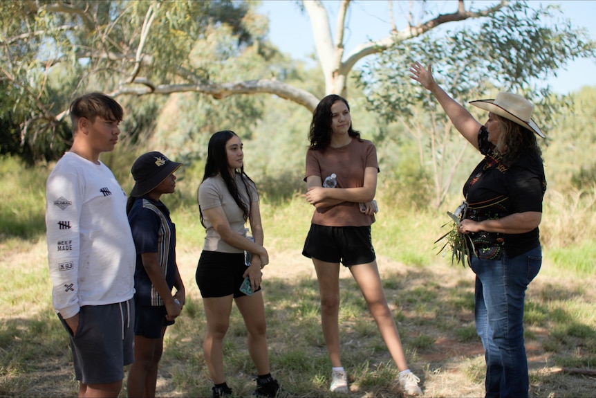 A woman stands with four younsters looking and listening to her in bush, blue skies.