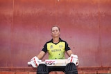 Female cricketer sitting in front of a painted wall with her bat and gloves