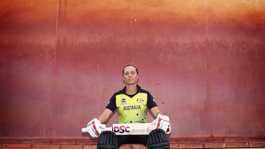Female cricketer sitting in front of a painted wall with her bat and gloves