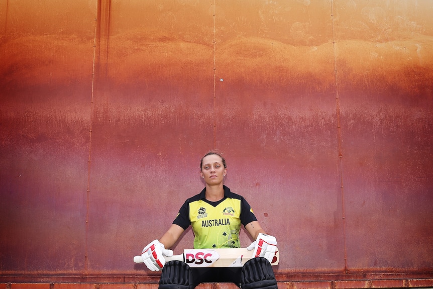 Female cricketer sitting in front of a painted wall with her bat and gloves