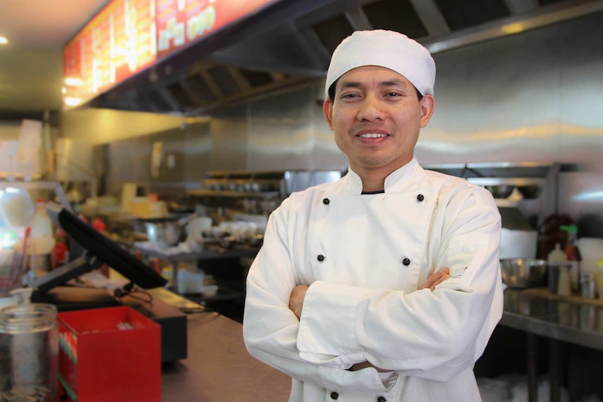 Man stands with arms folded wearing chef's uniform in industrial kitchen.