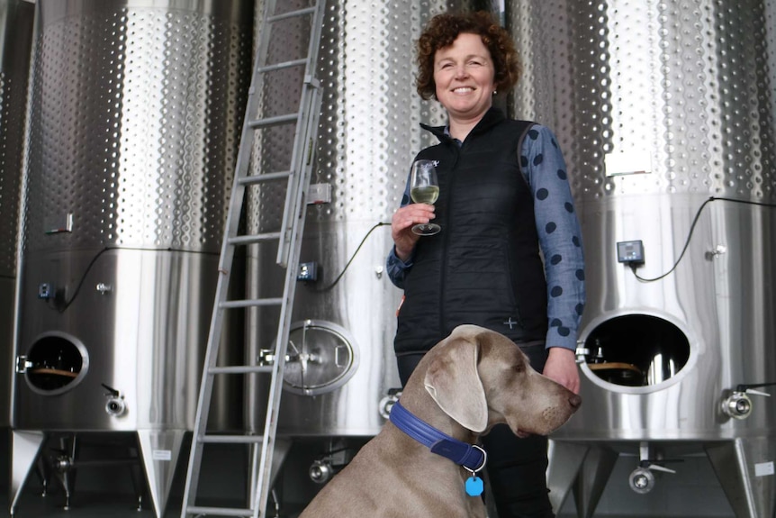 a dog stands next to winery owner in front of tall stainless steel wine tanks