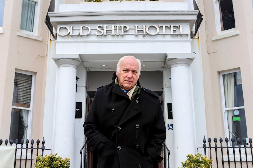 A white-haired man in an overcoat stands in front of the entrance to a building. Sign over door reads: Old Ship Hotel.