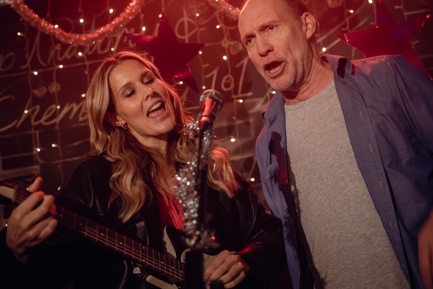 A middle-aged man and a young woman sing into a microphone decorated with tinsel.