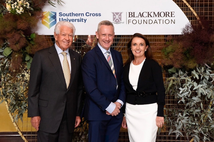 Two men in formal suits and a well dressed woman stand in front of a sign in a garden setting