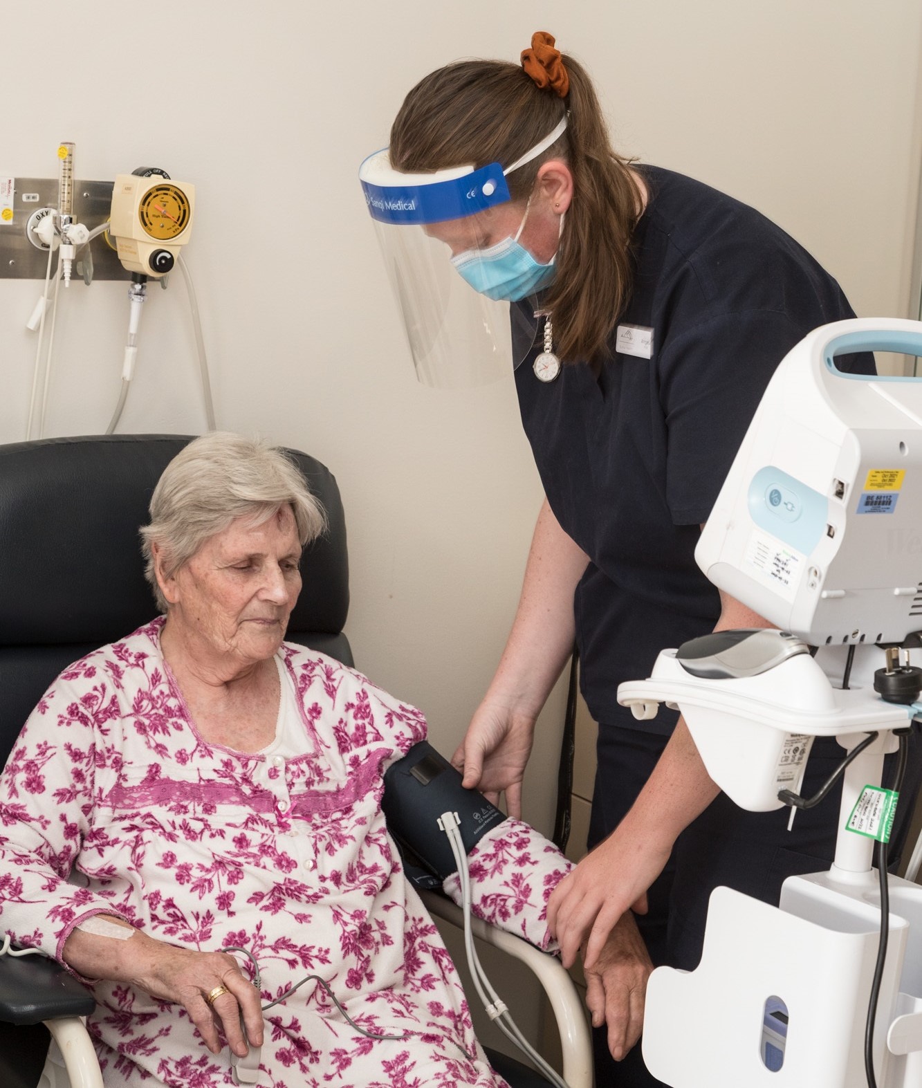 A nurse in a blue uniform and facemask and shield applies a blood pressure cuff to an older woman in a white and pink dress