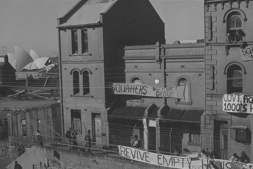 A row of buildings with protest signs including one that reads 'Revive Empty Houses' with the Opera House in the background.