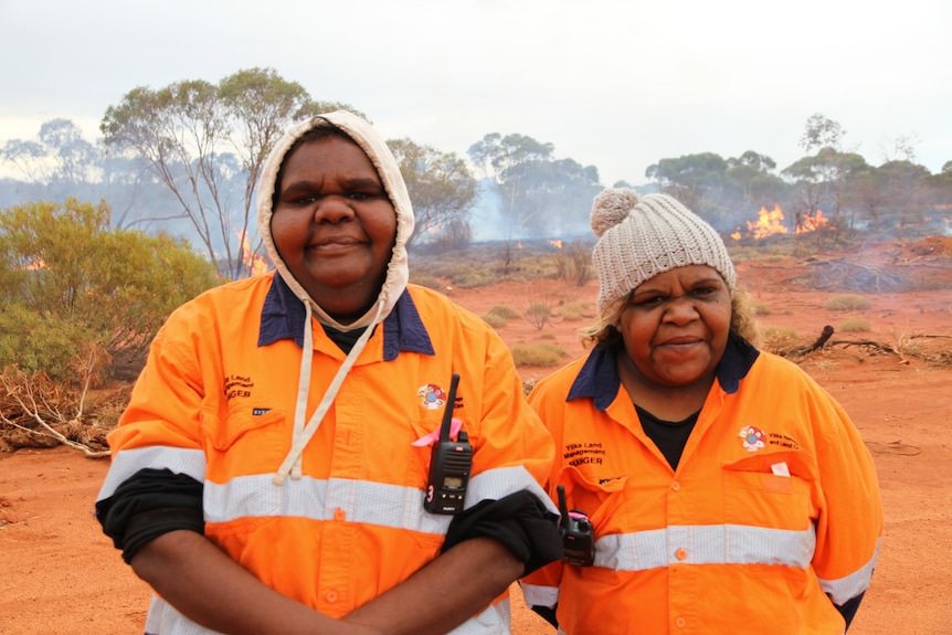 Two women stand next to each other smiling on a bare, flat patch of land