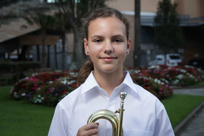 A 12-year-old girl in a white shirt stands in front a flower bed, holding a bugle.