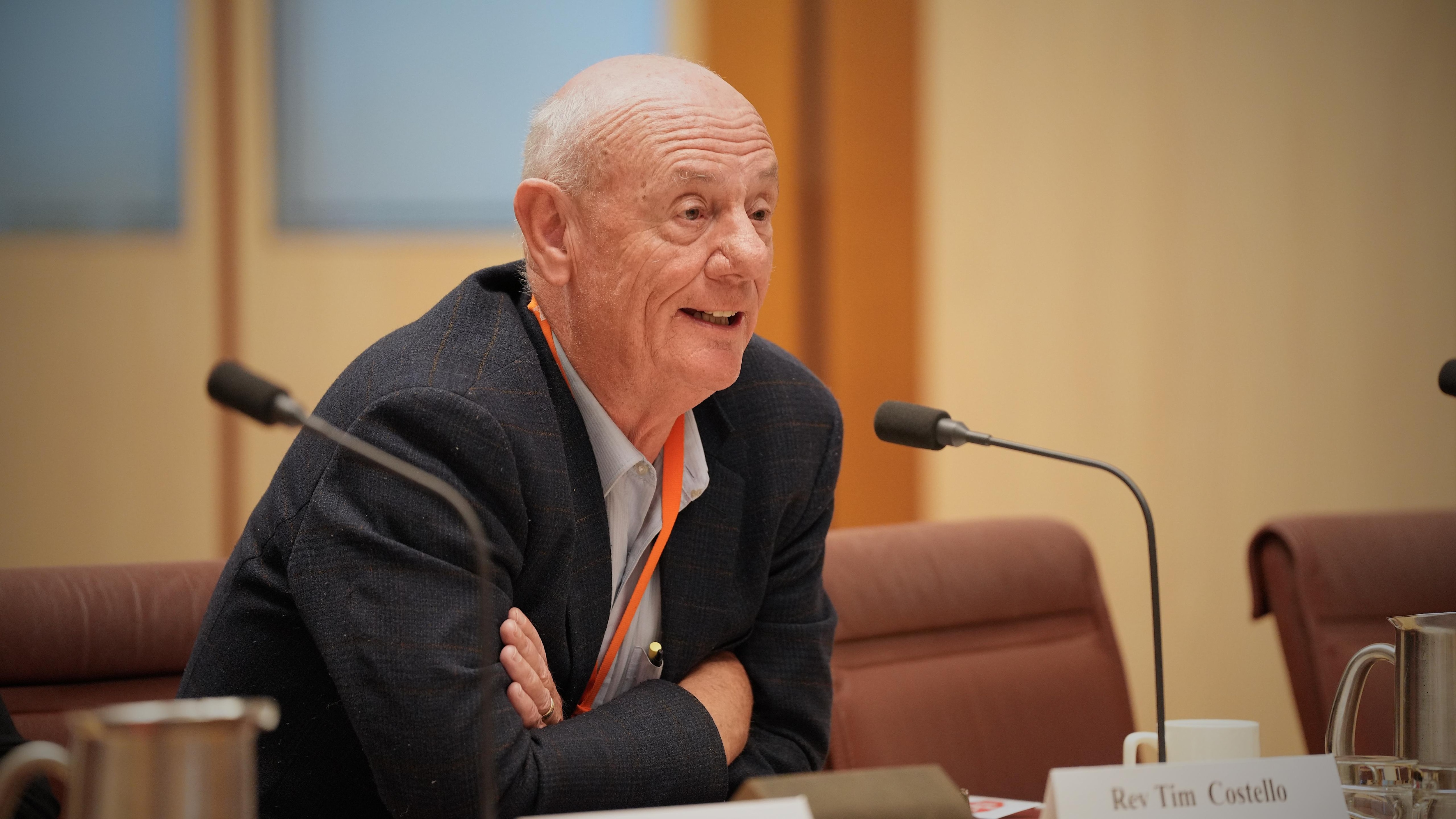 A man in a suit sits leaning forward on a desk and speaking into a microphone.