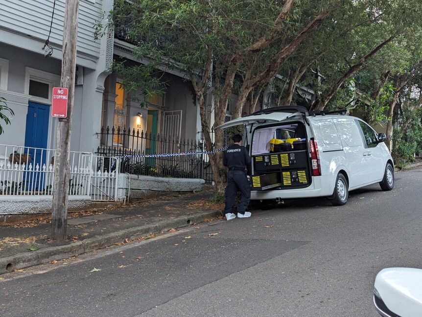 Un oficial de policía forense saca equipo de la parte trasera de una camioneta que está al lado de una casa con cinta policial.