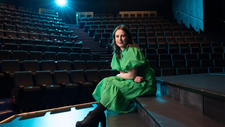 Laura Murphy, a brunette white woman in her 30s, wears a bright green dress and sits on a set piece on a stage in a dark theatre