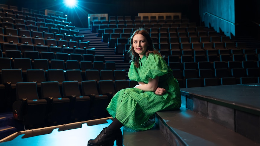 Laura Murphy, a brunette white woman in her 30s, wears a bright green dress and sits on a set piece on a stage in a dark theatre