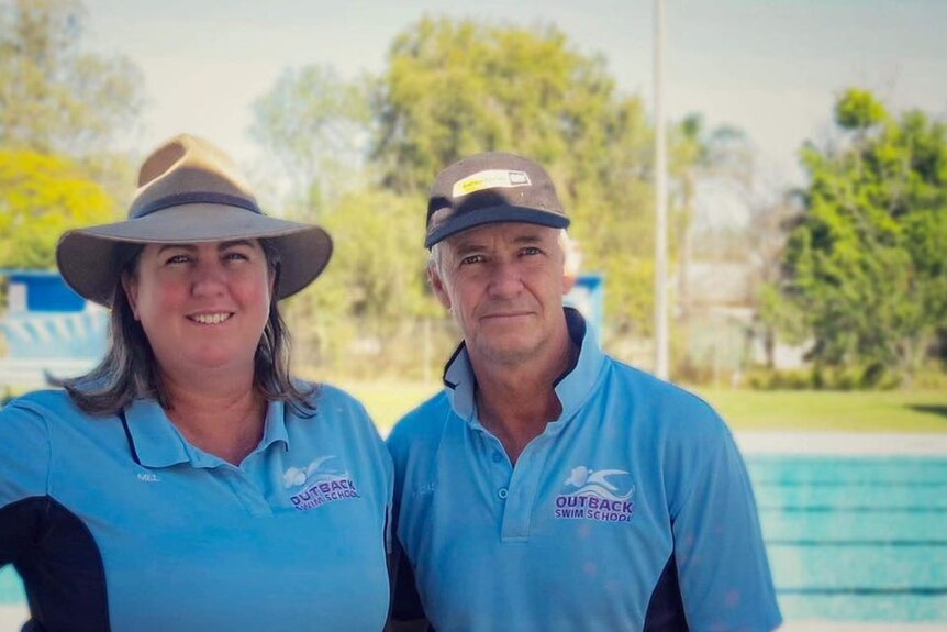 A woman and a man wearing matching polo shirts and hats stand in front of a pool, smiling.