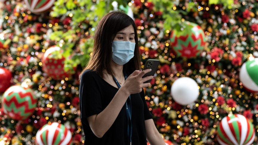 A woman wearing a mask looks at her mobile phone while in front of a colourful Christmas tree display.