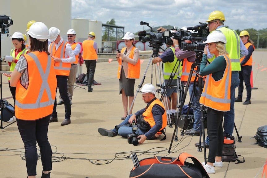 People stand around with cameras wearing hard hats and hi-vis vests.